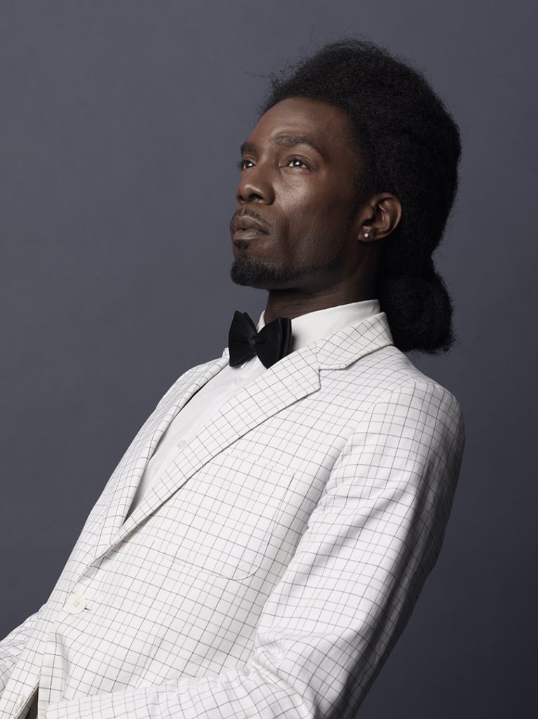 A portrait of a black man in a 2 piece white suit with a black bowtie.