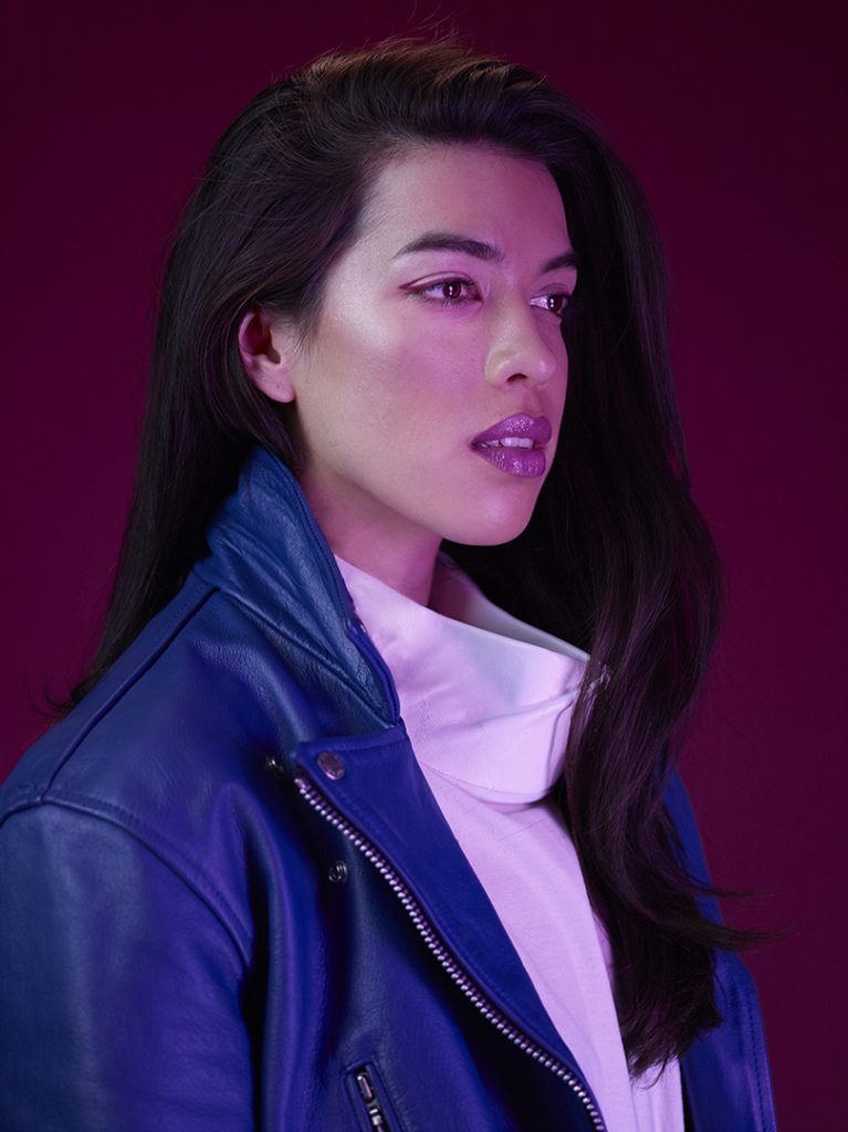 A studio portrait of a woman wearing a white top and blue jacket with a pink background.