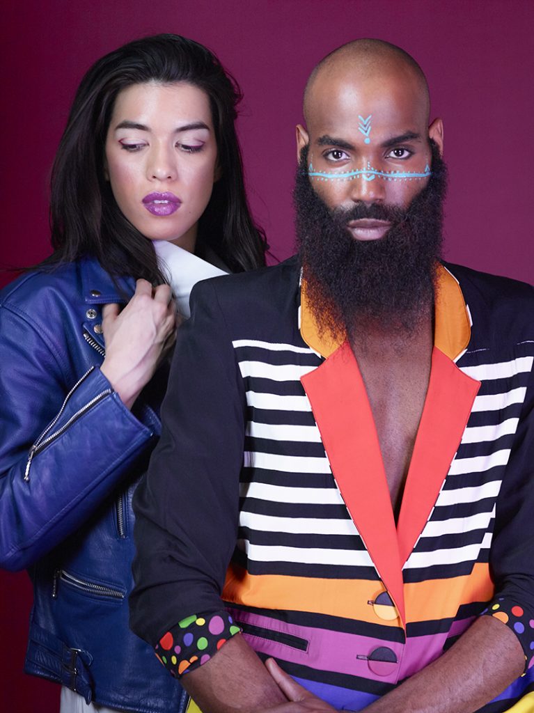 A studio portrait of a couple wearing vibrant clothing with a pink background.